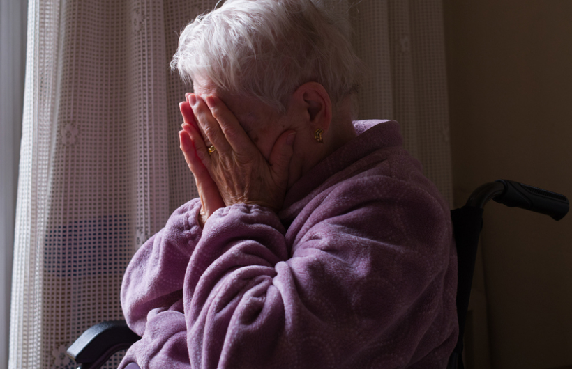 An elderly woman wearing a lavender sweatshirt covers her face with her hands while sitting by a window in her wheelchair