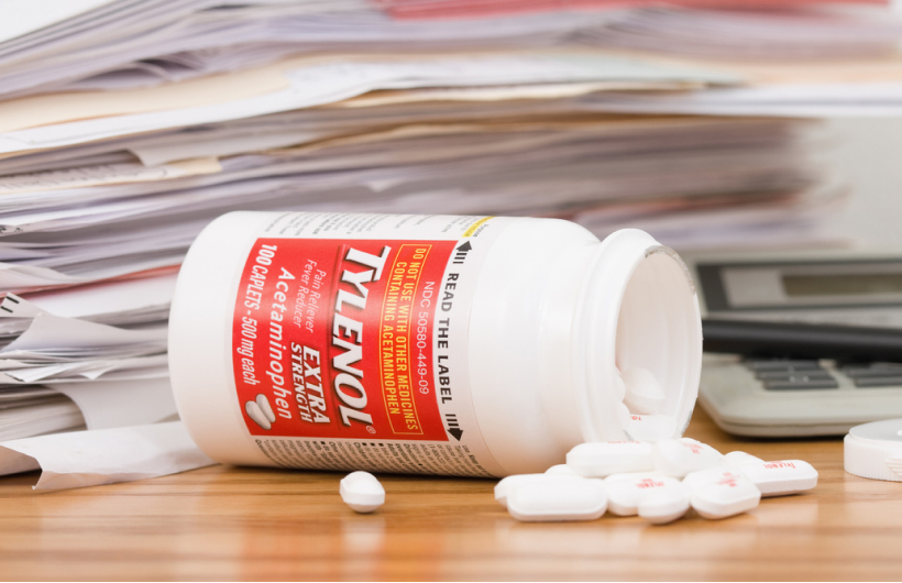 A white and red bottle of Tylenol sits on its side with the lid off. White Tylenol pills sit around it. A pile of white paperwork is in the background.