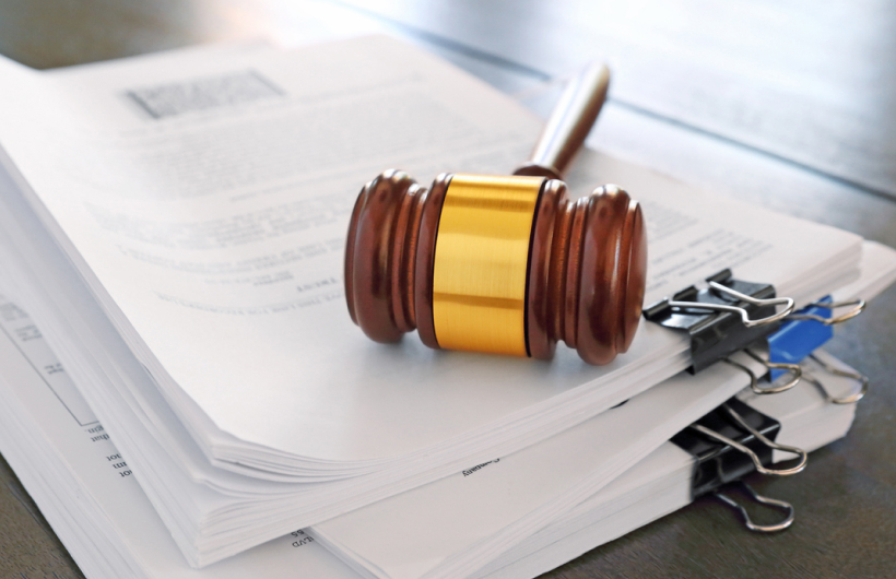 A brown wooden gavel with a gold band sits on a pile of sorted paperwork on a brown desk.