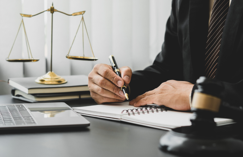 A person writes in a spiral-bound notebook with a black pen on a black desk. Golden scales of justice sit on stacked notebooks behind the person, who is wearing a black judge's robe.