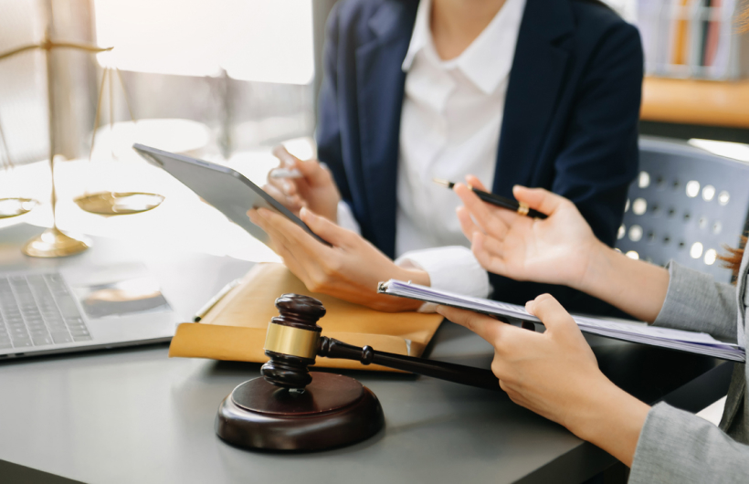 A woman in a navy blazer and white shirt sits at a desk with someone in a gray suit jacket. They discuss what's on their tablets while a brown wooden gavel sits in front of them.