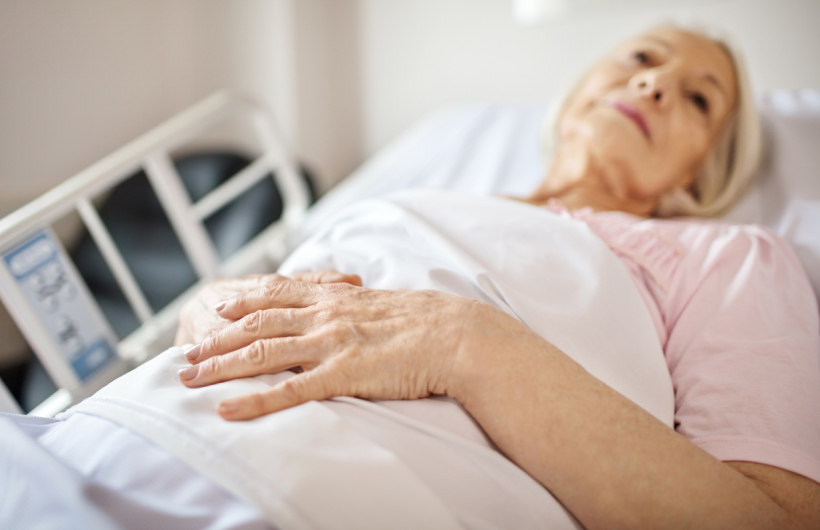 An elderly woman with white hair and a pink shirt lies in a hospital bed. She holds a white blanket over her chest and a black chair sits in the background.