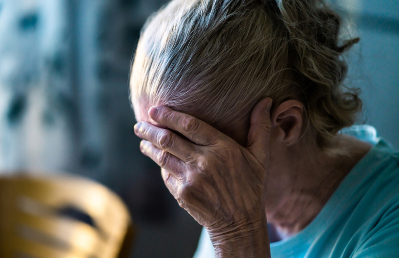 An older woman with gray hair and a teal shirt holds her face in her hand. A wooden chair and blue floral curtains are behind her.