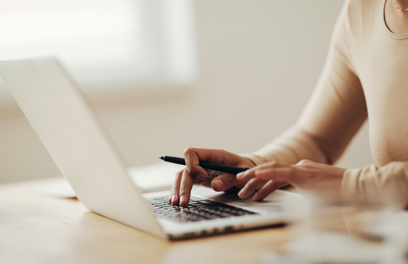 A woman in a tan shirt types on a Macbook while holding a pen. She sits at a light brown table in front of a sunny white window.