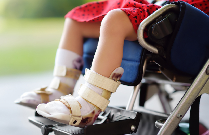 A young child sits in a wheelchair and wears special strapped shoes. They're sitting outside with green grass in the background.