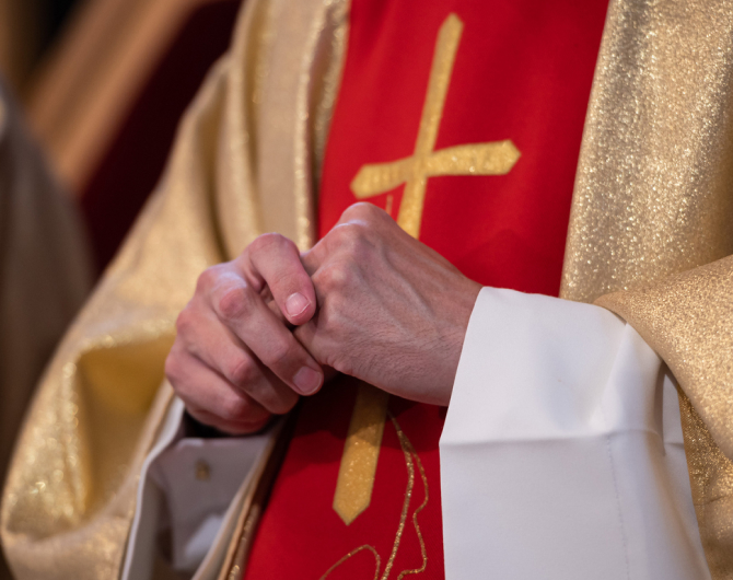 A church leader wearing a white and gold robe with a gold cross embroidered on a red sash stands with his hands together.