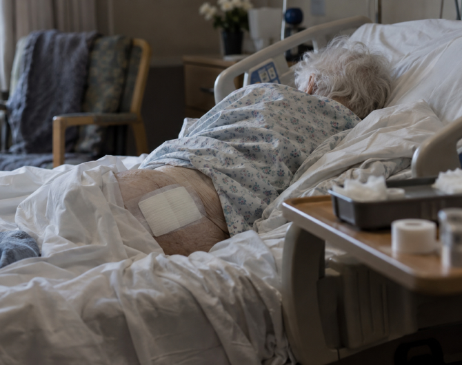 An older woman in a light blue hospital gown lays on her side in a hospital bed. She has a large bedsore bandage on her lower back.
